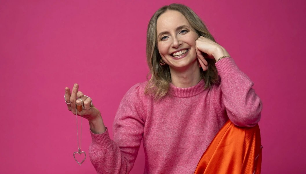 Katarina Blom i rosa tröja mot rosa bakgrund, leende med ett hjärthalsband i handen